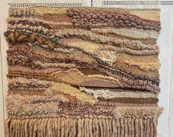 May include: A woven wall hanging in shades of beige, brown, and mauve. The textile features a variety of textures, including thick yarn, braids, and fringe. The piece is suspended from a wooden dowel with black knobs.