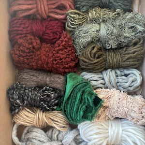 May include: A variety of colorful yarn and ribbon spools are arranged inside a cardboard box. The spools are in shades of red, brown, green, gray, and cream. The textures vary from smooth to textured, and the materials appear to be wool, cotton, and jute.