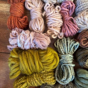 Pode incluir: Uma coleção de novelos de fios coloridos em vários tons de laranja, rosa, verde e bege. O fio é disposto sobre uma superfície de madeira, mostrando diferentes texturas e espessuras, ideal para projetos de artesanato.