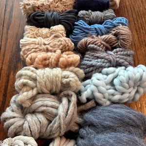 Könnte beinhalten: Eine Sammlung verschiedener Garnbündel in einer Reihe von Farben, darunter Beige, Grau, Blau und Braun. Die Garne sind auf einer Holzoberfläche angeordnet und zeigen verschiedene Texturen und Stärken, geeignet zum Stricken oder für andere Handarbeiten.