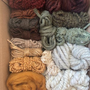 Puede incluir: Una variedad de ovillos y madejas de hilo de colores en una caja de cartón. Los hilos son en tonos de rojo, marrón, verde, beige y blanco. Algunos están bien enrollados, otros sueltos y texturizados, perfectos para proyectos de manualidades.
