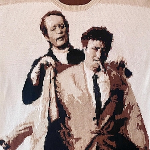 May include: Cream-colored knitted sweater featuring a two-person portrait in shades of brown, black, and cream. The design depicts two men in suits, with one draped over the other's shoulder. The sweater has a ribbed hem and neckline.