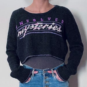 Può includere: Un maglione nero corto con una grafica "UNSOLVED MYSTERIES" in viola e rosa. Il maglione ha maniche lunghe e un collo a costine.