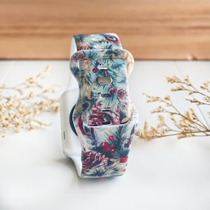 May include: A white watch band with a Christmas-themed print featuring pine branches, pine cones, berries, and gold lettering. The band is made of silicone and has a buckle closure.