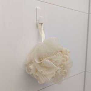 Op de afbeelding: Een witte badspons die aan een witte haak aan een witte betegelde muur hangt.