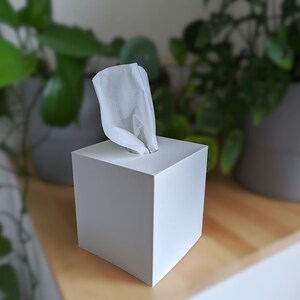 Könnte beinhalten: Eine weiße quadratische Taschentuchbox mit einem herausgezogenen weißen Taschentuch. Die Taschentuchbox steht auf einer Holzoberfläche.