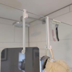 Könnte beinhalten: Weißer Dusch-Organizer für die Tür mit mehreren Haken. Der Organizer hängt an einer Glas-Duschwand und hält einen Luffa, eine Bürste und anderes Duschzubehör. Der Duschkopf ist im Hintergrund sichtbar.