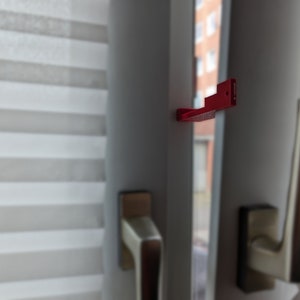 Pode incluir: Um fecho de janela de plástico vermelho com um pequeno furo no centro. O fecho está preso a uma moldura de janela branca.