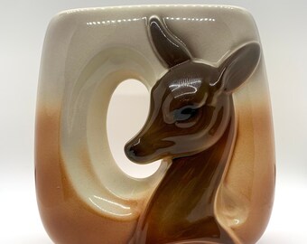 May include: A ceramic vase with a brown and white glaze features a deer head design. The deer's head is in relief and has a brown glaze. The vase has a rounded shape and a wide opening.