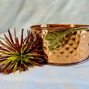 May include: A hammered copper bowl with a brass vine and leaf design. The bowl has a rounded shape and a smooth, polished finish.