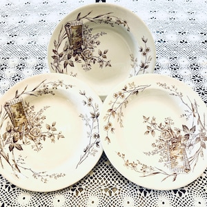 Pode incluir: Três pratos de cerâmica creme com um desenho floral castanho. Cada prato apresenta uma imagem central de uma moldura e flores. Os pratos estão dispostos sobre uma toalha de mesa de renda branca.