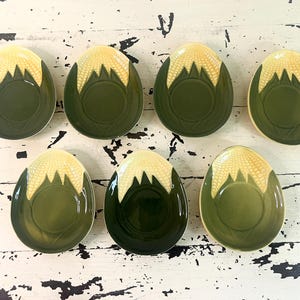 Peut inclure: Sept petites assiettes en céramique vert olive en forme d'œuf, avec un motif texturé de couleur crème en haut. Les assiettes sont disposées sur une surface en bois blanc vieilli. Le motif ressemble à un épi de maïs.