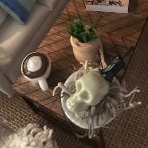 May include: Miniature living room scene with a white sofa, blue striped pillow, and a wooden coffee table. The table holds a small orange pumpkin, a book, a potted plant, a coffee cup, and a skull decoration, suggesting a Halloween theme.
