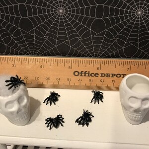 Puede incluir: Dos platos de caramelo de calavera de cer&aacute;mica blanca con ara&ntilde;as de pl&aacute;stico negro encima y alrededor de ellos. Las calaveras est&aacute;n sentadas en una mesa blanca con una regla de madera detr&aacute;s de ellas. La regla tiene las palabras "Office Depot" impresas en ella.