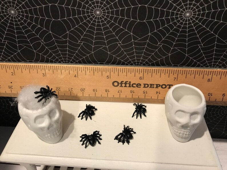 Puede incluir: Dos platos de caramelo de calavera de cer&aacute;mica blanca con ara&ntilde;as de pl&aacute;stico negro encima y alrededor de ellos. Las calaveras est&aacute;n sentadas en una mesa blanca con una regla de madera detr&aacute;s de ellas. La regla tiene las palabras "Office Depot" impresas en ella.