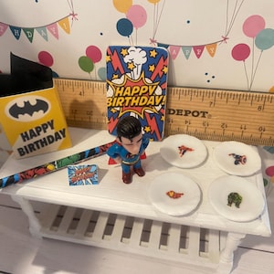 May include: A miniature birthday party scene with superhero-themed decorations. Includes a Superman figurine, small plates with superhero logos, a Batman-themed gift box, and a "Happy Birthday" card. The background features a festive banner.