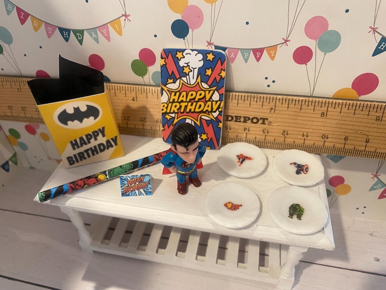 May include: A miniature birthday party scene with superhero-themed decorations. Includes a Superman figurine, small plates with superhero logos, a Batman-themed gift box, and a "Happy Birthday" card. The background features a festive banner.