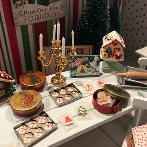 Puede incluir: Una escena navideña en miniatura con una casa de jengibre, un rodillo y varias galletas navideñas en latas y en bandejas para hornear. La escena incluye un candelabro dorado con velas blancas, un árbol de Navidad y un letrero que dice "All Hearts Come Home For Christmas".