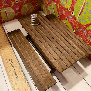 May include: A small wooden picnic table with a thimble on top. The table is made of dark brown wood and has a slatted design. A wooden ruler is next to the table. The background features a colorful fabric with a repeating pattern.