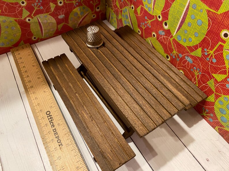 May include: A small wooden picnic table with a thimble on top. The table is made of dark brown wood and has a slatted design. A wooden ruler is next to the table. The background features a colorful fabric with a repeating pattern.