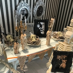 May include: A miniature Halloween scene with skeletons, skulls, and candles on a white table. The table is decorated with a burlap banner and framed vintage photos. The background features black and white striped wallpaper and framed portraits.