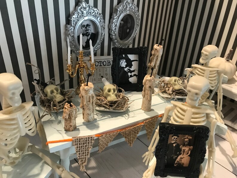 May include: A miniature Halloween scene with skeletons, skulls, and candles on a white table. The table is decorated with a burlap banner and framed vintage photos. The background features black and white striped wallpaper and framed portraits.