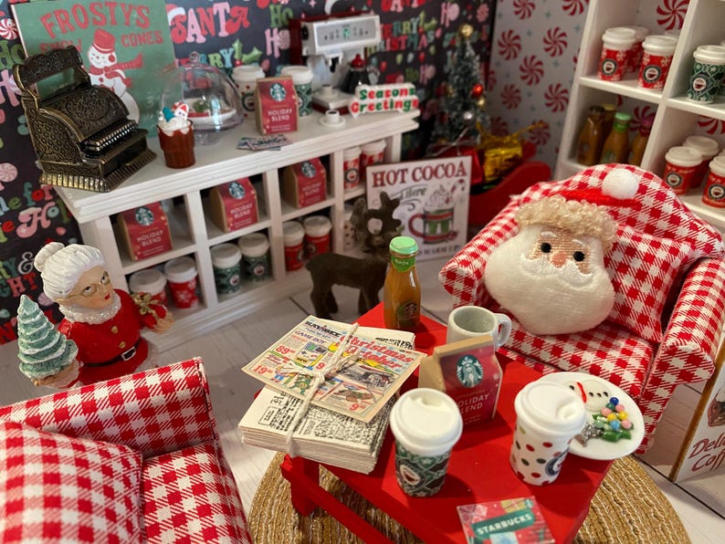 Pode incluir: Uma loja de Natal em miniatura com uma poltrona xadrez vermelha e branca, uma mesa vermelha com uma pilha de jornais e uma boneca do Papai Noel. A loja tem um balc&atilde;o branco com prateleiras cheias de itens de Natal em miniatura, incluindo x&iacute;caras de caf&eacute;, bengalas de doces e um caixa registradora.