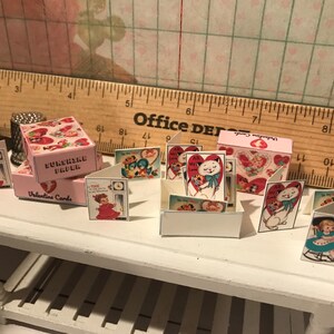 Może przedstawiać: Miniaturowe kartki i pudełka walentynkowe. Kartki przedstawiają ilustracje w stylu vintage kot&oacute;w, dzieci i serc. Pudełka są r&oacute;żowe i mają napisy "Sunshine Paper" i "Valentine Cards". Kartki są wystawione na białym stole.