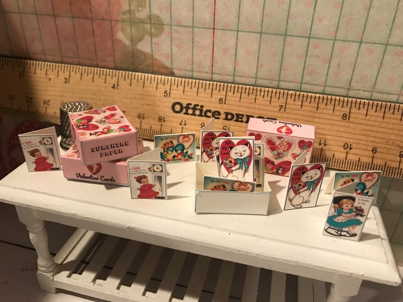 Może przedstawiać: Miniaturowe kartki i pudełka walentynkowe. Kartki przedstawiają ilustracje w stylu vintage kot&oacute;w, dzieci i serc. Pudełka są r&oacute;żowe i mają napisy "Sunshine Paper" i "Valentine Cards". Kartki są wystawione na białym stole.