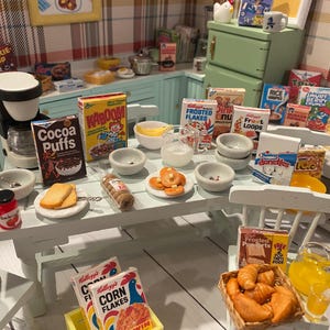 May include: A miniature kitchen scene with a white table set with cereal boxes, bowls, and food items. Boxes of Cocoa Puffs, Kaboom, and Frosted Flakes are visible. A coffee maker and a jug of juice are also present. The scene is set in a dollhouse.
