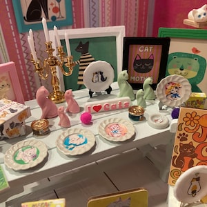 K&ouml;nnte beinhalten: Ein wei&szlig;er Miniaturtisch mit verschiedenen katzenbezogenen Gegenst&auml;nden, darunter kleine dekorative Teller mit Illustrationen, ein goldener Kandelaber, rosa Katzenfiguren, ein Schild mit der Aufschrift I Love Cats und Bilderrahmen.