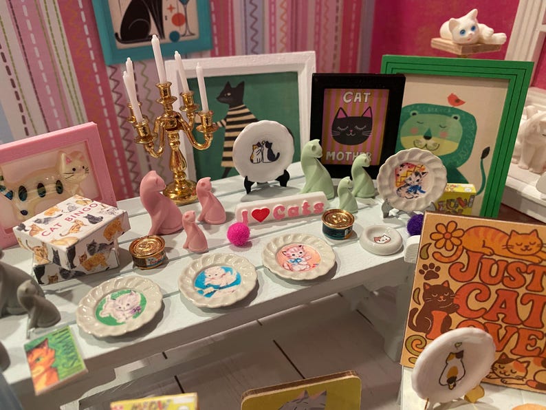 K&ouml;nnte beinhalten: Ein wei&szlig;er Miniaturtisch mit verschiedenen katzenbezogenen Gegenst&auml;nden, darunter kleine dekorative Teller mit Illustrationen, ein goldener Kandelaber, rosa Katzenfiguren, ein Schild mit der Aufschrift I Love Cats und Bilderrahmen.