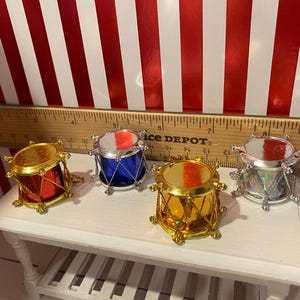 May include: Four miniature toy drums in red, blue, gold, and silver, each with a metallic finish and a small size. The drums are arranged on a white surface, with a ruler in the background. The background has red and white stripes.
