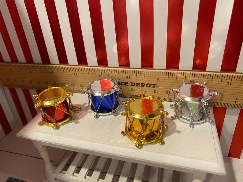 May include: Four miniature toy drums in red, blue, gold, and silver, each with a metallic finish and a small size. The drums are arranged on a white surface, with a ruler in the background. The background has red and white stripes.