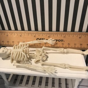 May include: A small, off-white plastic skeleton figurine, lying on a white table. A wooden ruler with black markings is placed above the skeleton. The background features a black and white striped pattern.
