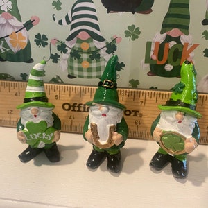 Puede incluir: Tres gnomos verdes con barbas blancas y labios rojos. Cada gnomo sostiene un amuleto de la suerte diferente: un tr&eacute;bol de cuatro hojas, una herradura y un tr&eacute;bol. Los gnomos est&aacute;n de pie sobre una superficie blanca.