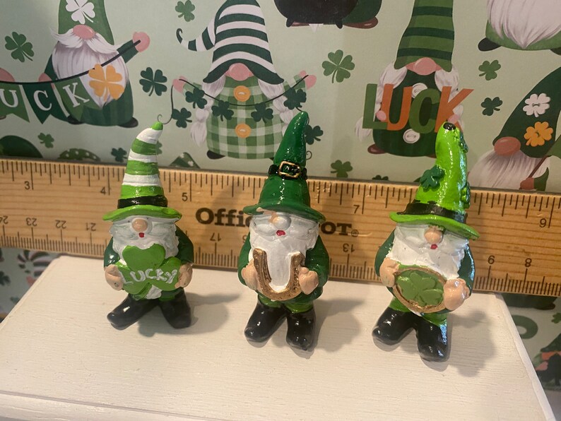 Puede incluir: Tres gnomos verdes con barbas blancas y labios rojos. Cada gnomo sostiene un amuleto de la suerte diferente: un tr&eacute;bol de cuatro hojas, una herradura y un tr&eacute;bol. Los gnomos est&aacute;n de pie sobre una superficie blanca.
