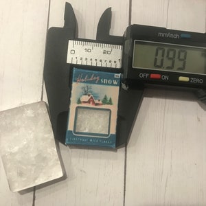 May include: A small, rectangular block of clear, crystalline material next to a miniature box labeled "Holiday Snow." The box, approximately 0.99 inches wide, features a winter scene with a red house and evergreen trees. A digital caliper measures the box's width.