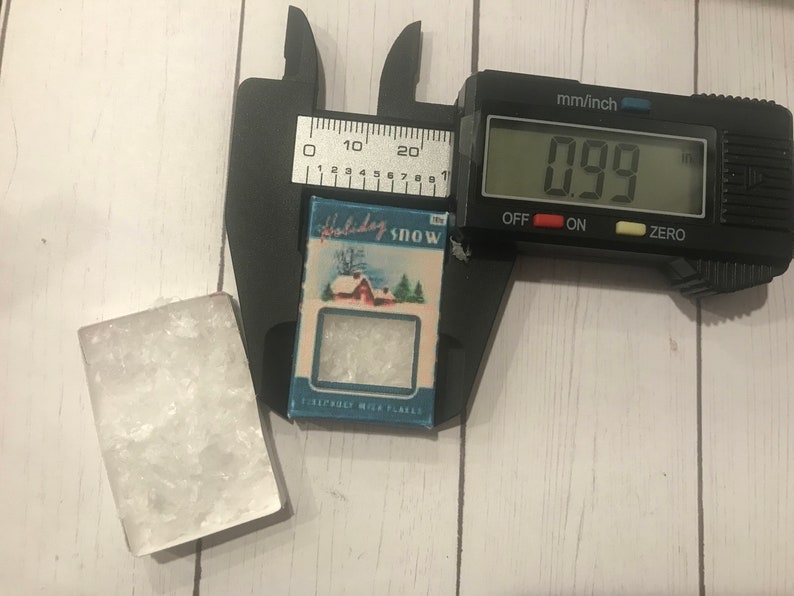 May include: A small, rectangular block of clear, crystalline material next to a miniature box labeled "Holiday Snow." The box, approximately 0.99 inches wide, features a winter scene with a red house and evergreen trees. A digital caliper measures the box's width.