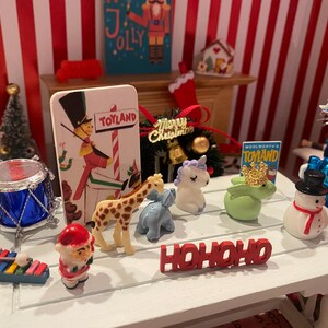 May include: A miniature Christmas scene with a white table displaying festive figurines. Includes a drum, a giraffe, a snowman, a unicorn, and a "HO HO HO" sign. A "Toyland" sign and a Christmas tree are also visible.