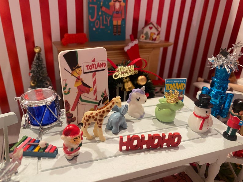 May include: A miniature Christmas scene with a white table displaying festive figurines. Includes a drum, a giraffe, a snowman, a unicorn, and a "HO HO HO" sign. A "Toyland" sign and a Christmas tree are also visible.