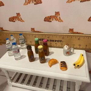 Pode incluir: Miniaturas de alimentos numa mesa branca, incluindo garrafas de &aacute;gua, bebidas de caf&eacute;, um croissant, um pastel, uma caneca de chantilly e bananas. O fundo apresenta um papel de parede com estampa de tigre e uma r&eacute;gua de madeira.