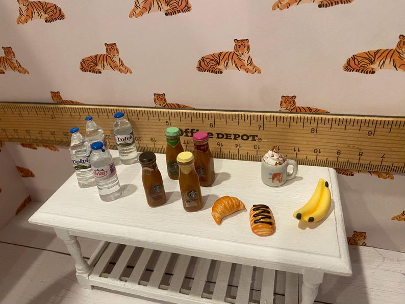 Pode incluir: Miniaturas de alimentos numa mesa branca, incluindo garrafas de &aacute;gua, bebidas de caf&eacute;, um croissant, um pastel, uma caneca de chantilly e bananas. O fundo apresenta um papel de parede com estampa de tigre e uma r&eacute;gua de madeira.
