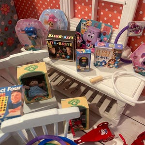 Op de afbeelding: Een verzameling miniatuurspeelgoed, waaronder een Lite-Brite-doos, een Cabbage Patch Kids-pop en een Connect Four-spel. Andere speelgoed zijn een paars personage, een hoelahoep en een pop in een roze verpakking. Het speelgoed is gerangschikt op een witte plank.