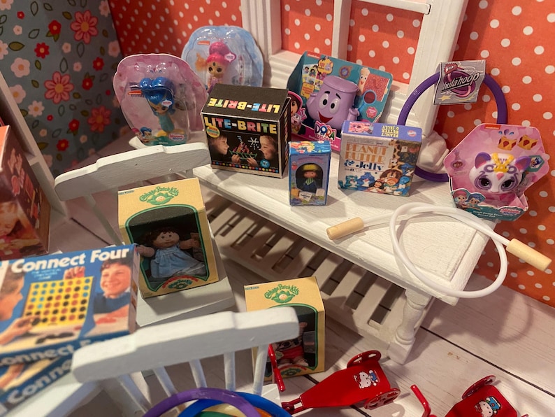 Op de afbeelding: Een verzameling miniatuurspeelgoed, waaronder een Lite-Brite-doos, een Cabbage Patch Kids-pop en een Connect Four-spel. Andere speelgoed zijn een paars personage, een hoelahoep en een pop in een roze verpakking. Het speelgoed is gerangschikt op een witte plank.