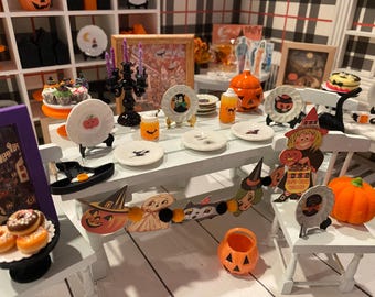 Könnte beinhalten: Ein Miniatur-Halloween-Esszimmerset mit weißem Tisch und Stühlen, dekoriert mit kleinen Tellern, einem schwarzen Kandelaber mit violetten Kerzen, orangefarbenen Kürbisbehältern, einer Girlande aus Papierfiguren und winzigen Lebensmitteln.