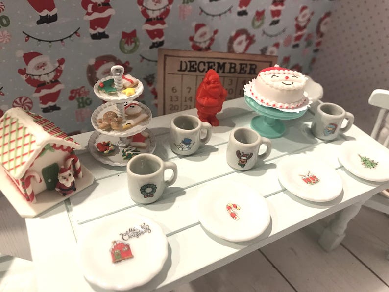 May include: A miniature Christmas scene with a gingerbread house, a tiered cake stand with Christmas cookies, and a white cake with a red Santa hat. There are also miniature mugs and plates with Christmas designs.