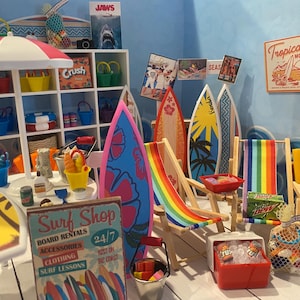 Peut inclure: Une scène de plage miniature avec des planches de surf, des chaises longues et un parasol à rayures rouges et blanches. La scène comprend une enseigne "Surf Shop", une enseigne "Tropicana Motel" et une affiche du film "JAWS", évoquant l'été.