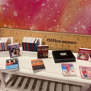 K&ouml;nnte beinhalten: Eine Sammlung von B&uuml;chern und DVDs mit beliebten Fernsehsendungen, darunter 'Friends', 'Clueless' und 'The O.C.', die auf einem wei&szlig;en Regal und Tisch ausgestellt sind.