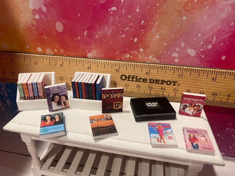 K&ouml;nnte beinhalten: Eine Sammlung von B&uuml;chern und DVDs mit beliebten Fernsehsendungen, darunter 'Friends', 'Clueless' und 'The O.C.', die auf einem wei&szlig;en Regal und Tisch ausgestellt sind.